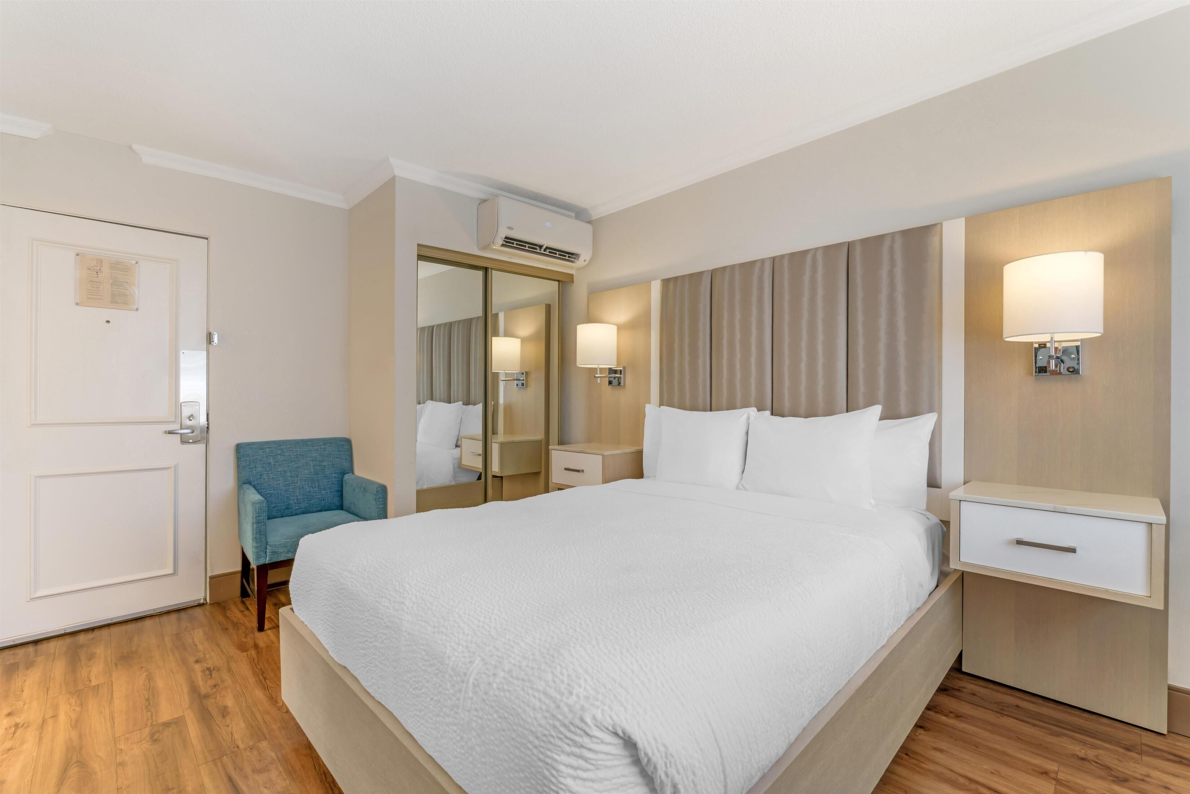 A hotel room with a large bed, a chair, a bedside table, a lamp, a mirror, and an air conditioning unit.