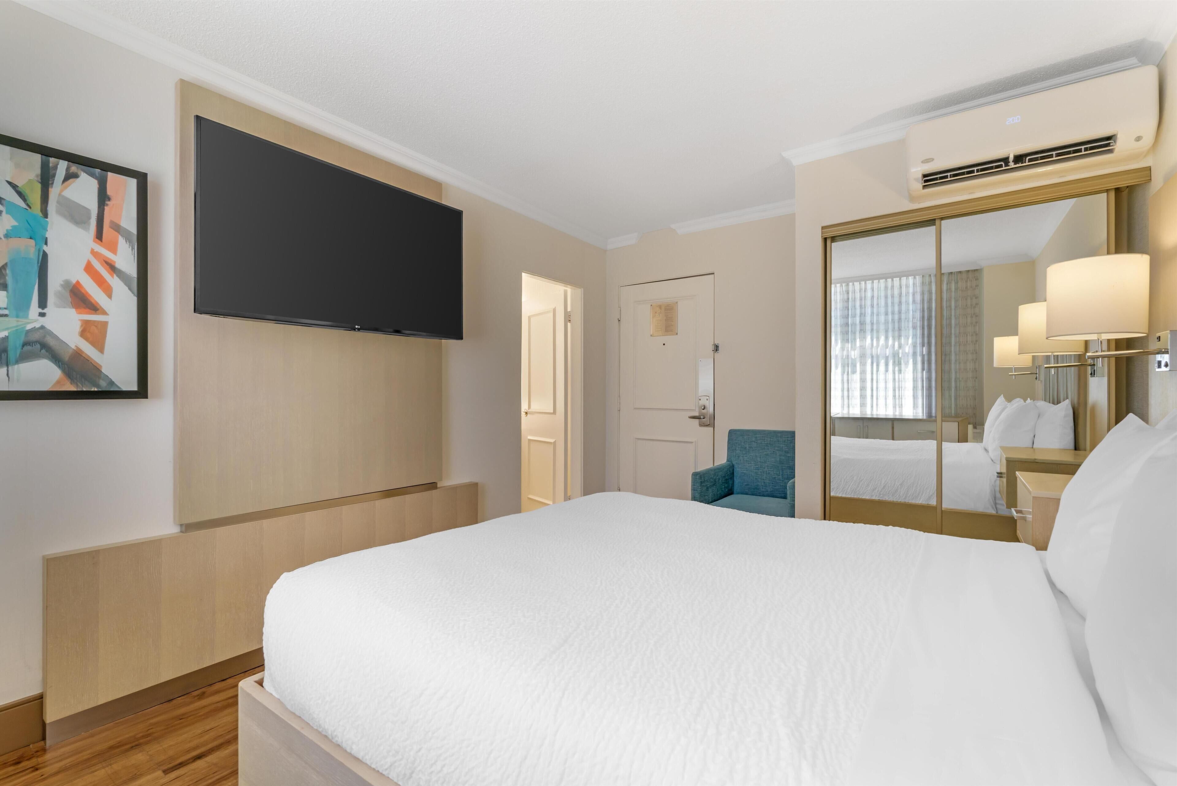 A hotel room with a bed, a flat-screen TV, a mirror, a chair, and a wardrobe.
