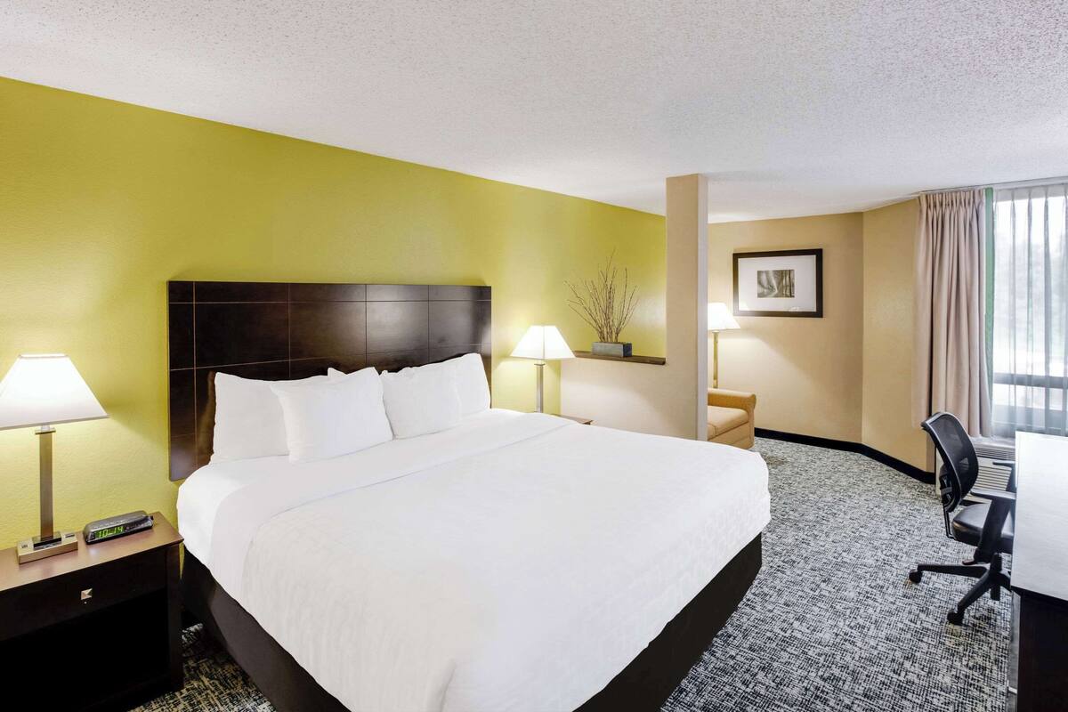 A hotel room with a large bed, two bedside tables with lamps, a desk with a chair, and a window with curtains.