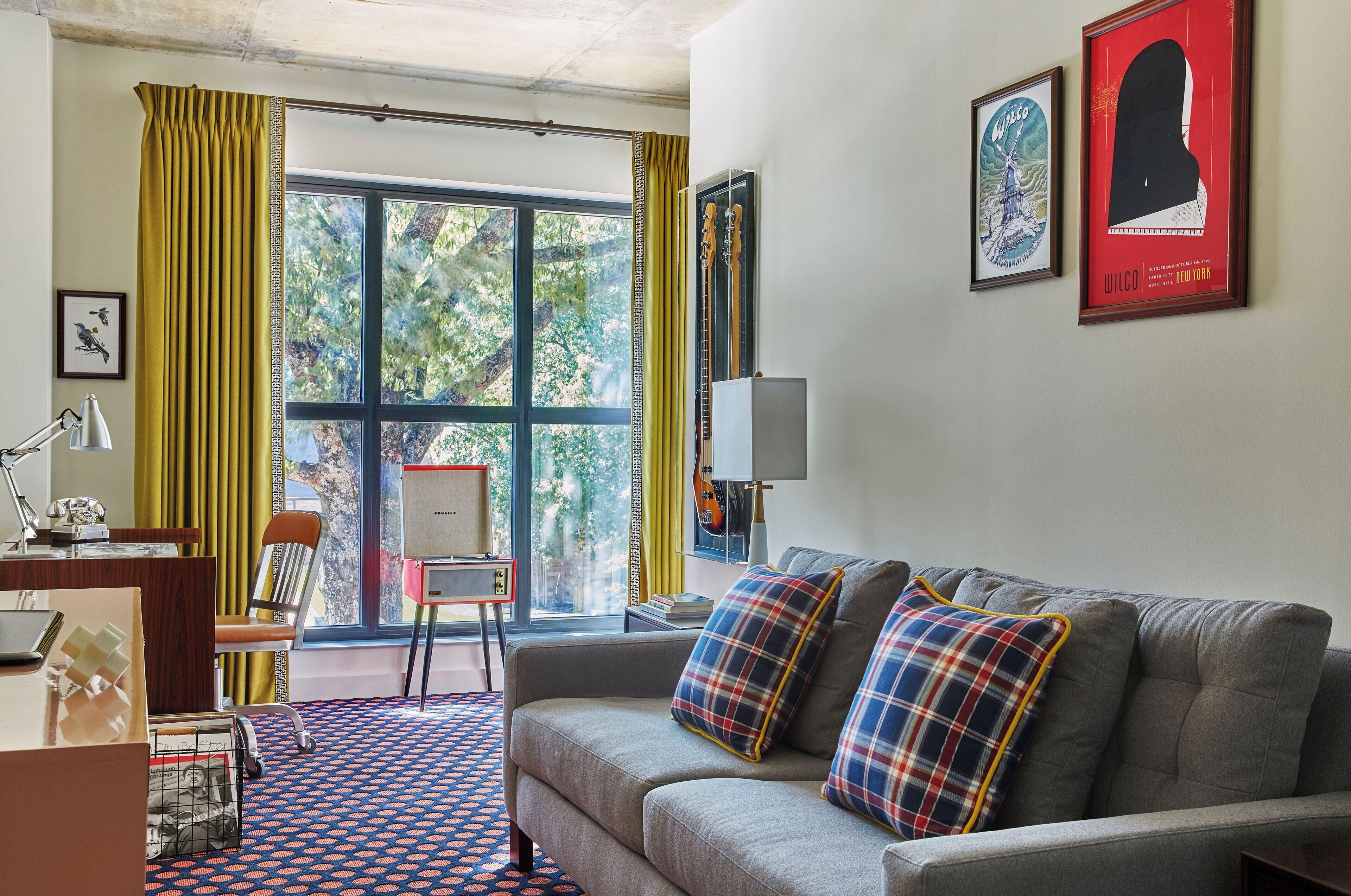 A hotel room with a large window, a sofa with plaid cushions, a desk with a lamp, and framed artwork on the wall.