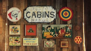 A cozy cabin-themed room with a wooden wall, a sofa, two chairs, and various wall decorations including a sign that reads "CABINS MANU".