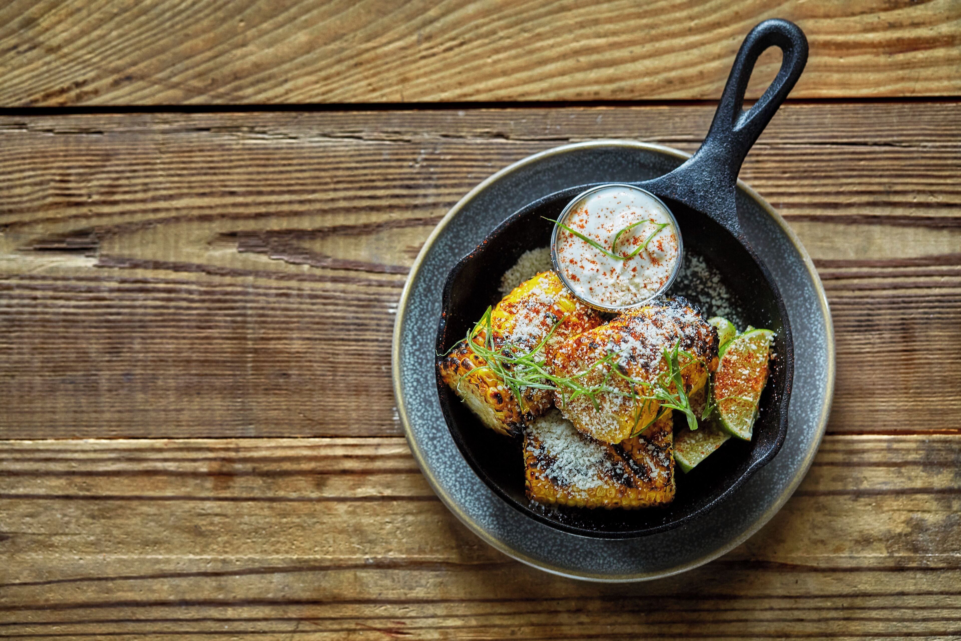 A skillet with grilled corn and lime wedges on a wooden surface.