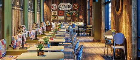 A long dining area with wooden walls and a sign reading "CABINS MANOR". Tables are set with checkered chairs and potted plants.