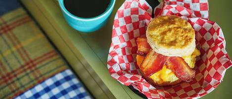 Un sándwich de desayuno en un recipiente de papel, una taza de café y una servilleta a cuadros sobre una mesa.