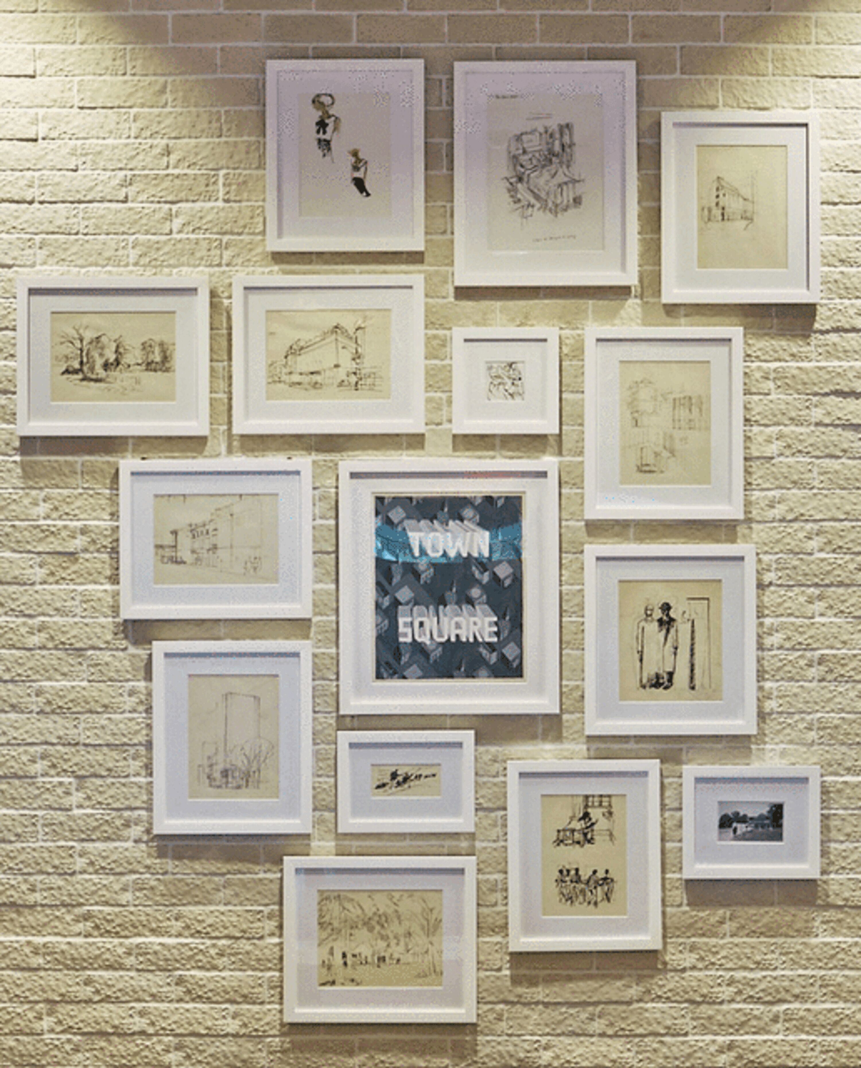 Una pared con múltiples bocetos y dibujos enmarcados, incluyendo uno que dice "TOWN SQUARE".