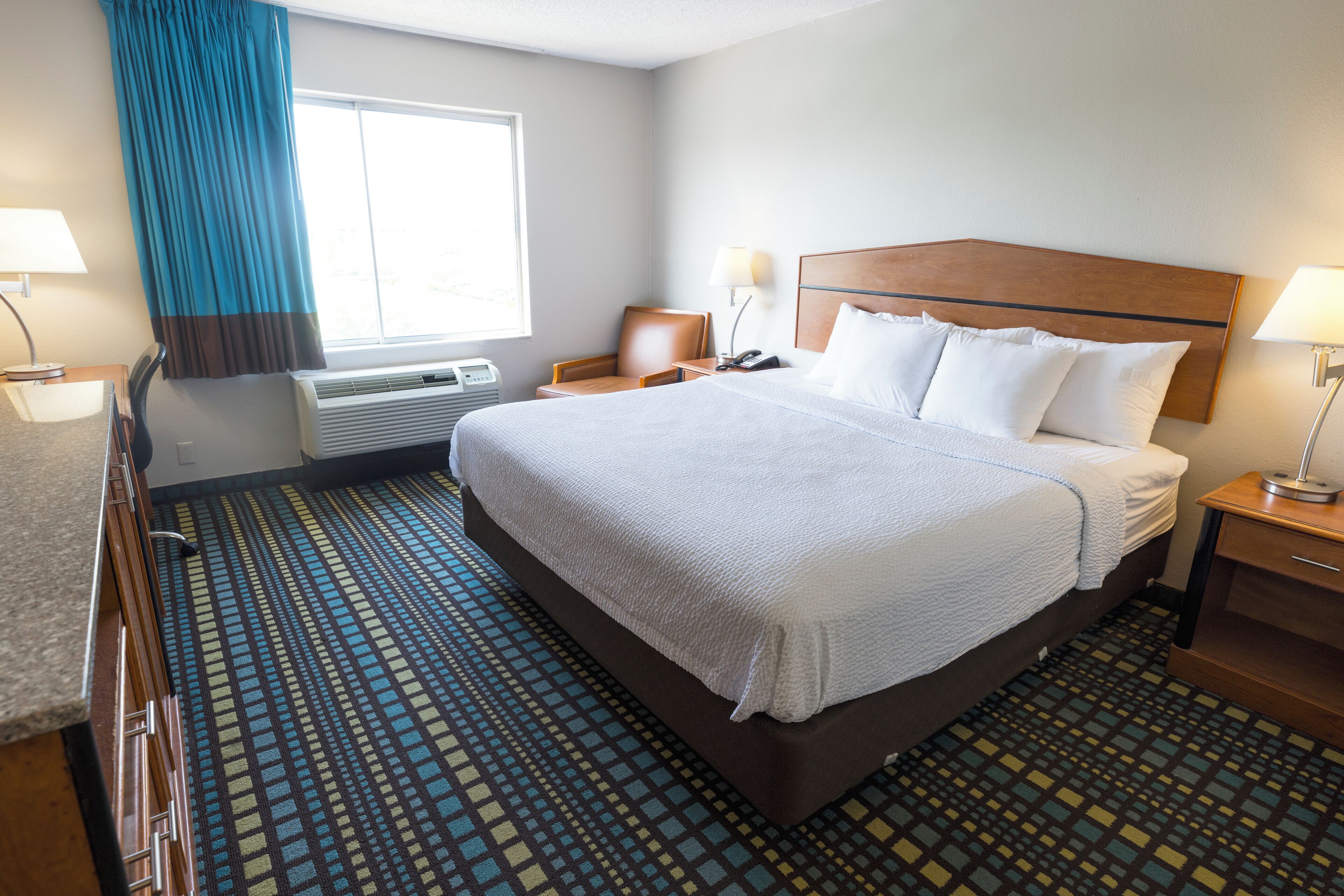 Quarto de hotel com uma cama grande, uma escrivaninha com luminária, uma cadeira e uma janela com cortinas.