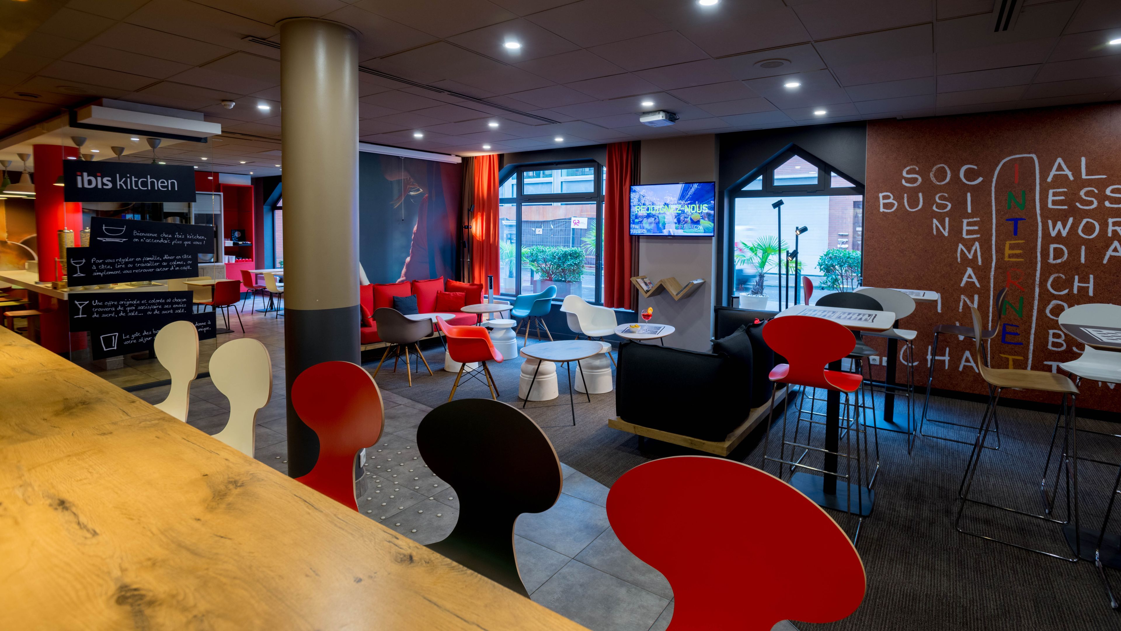 A modern restaurant interior with a mix of red and white chairs, a wooden counter, and a large window offering a view of the street.