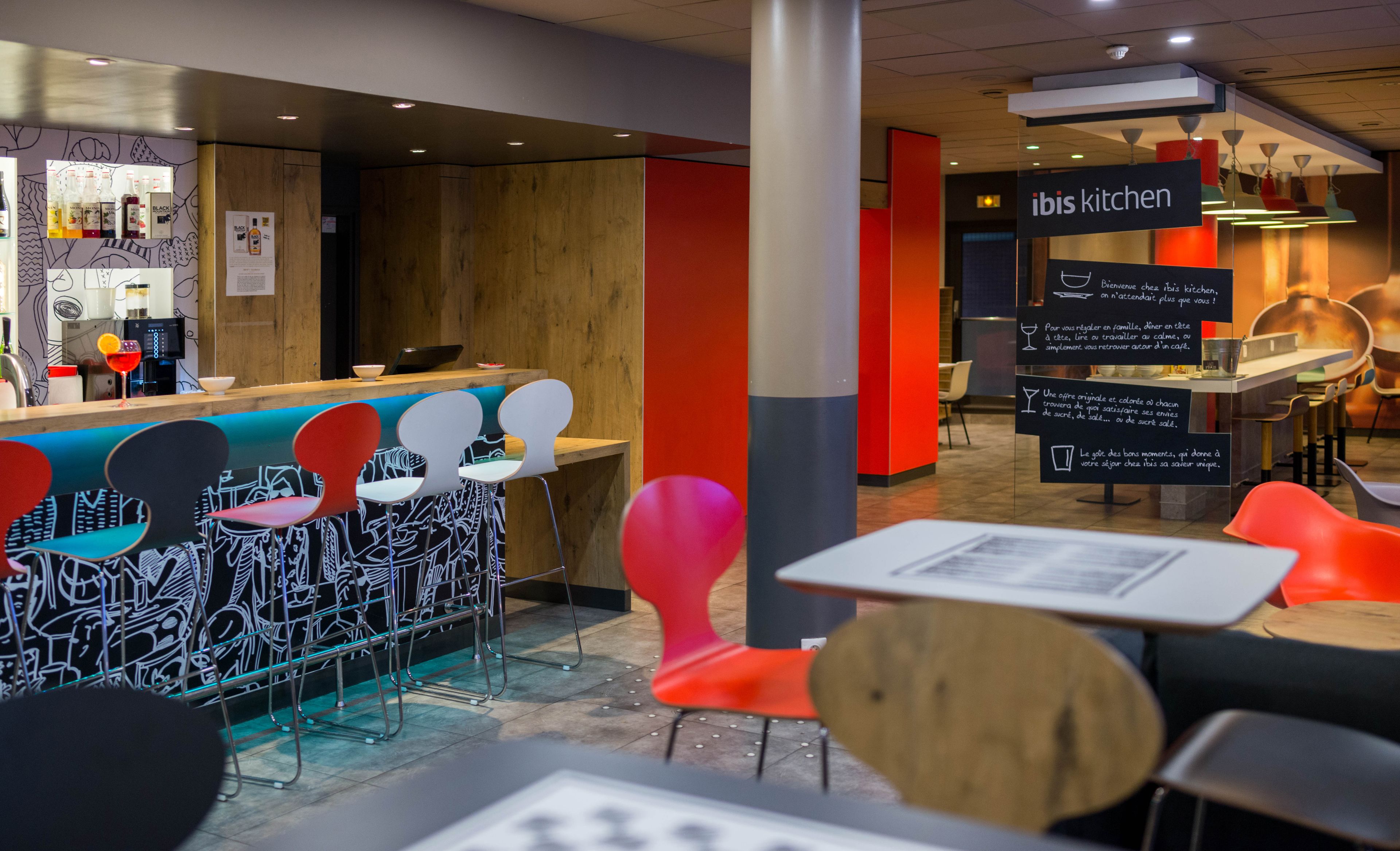 Zona de bar moderna con mostrador de madera, sillas coloridas y un letrero que indica 'ibis kitchen'.