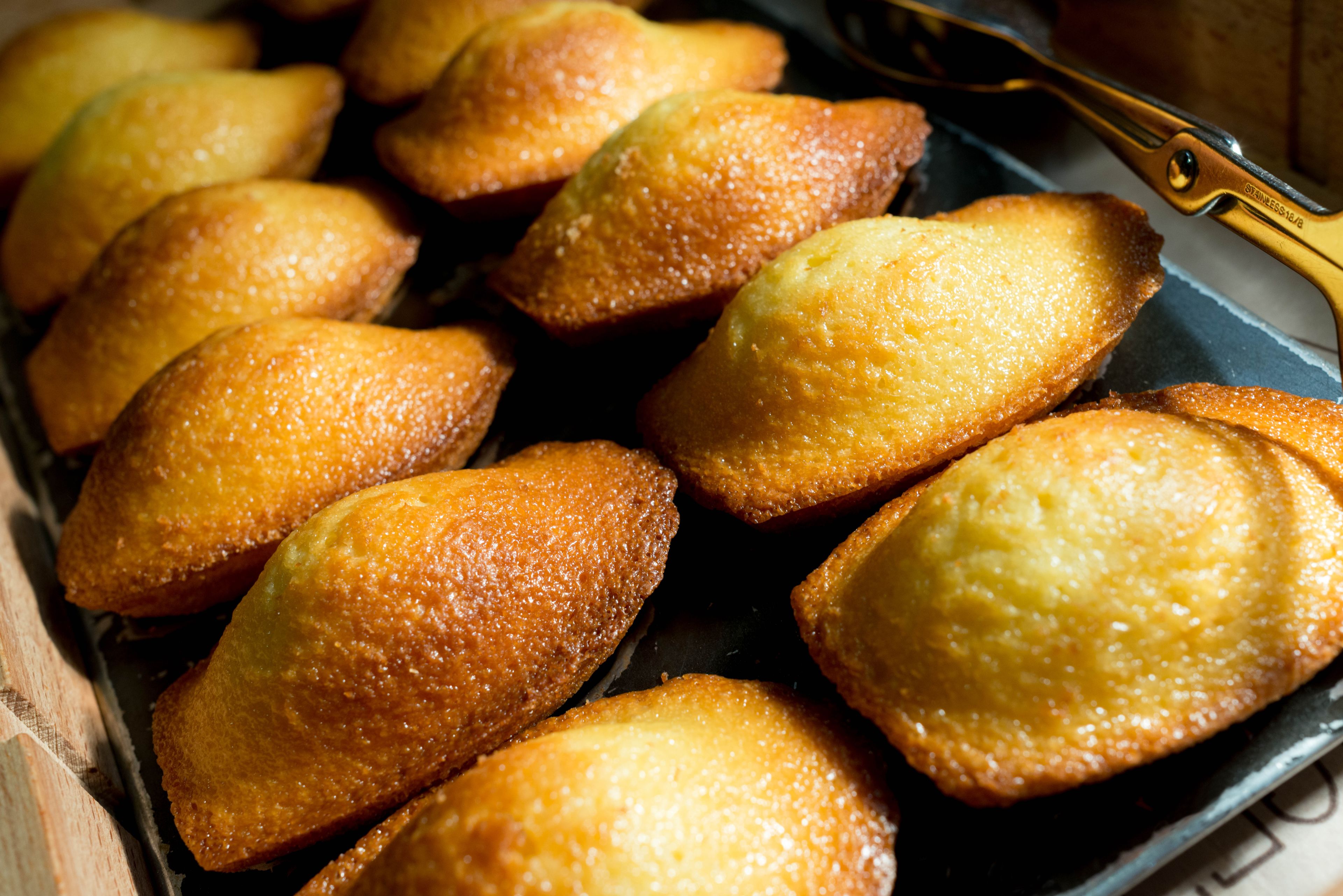 Una bandeja de pasteles dorados y con la superficie texturizada, probablemente espolvoreados con azúcar, acompañados de un par de pinzas con mangos dorados.