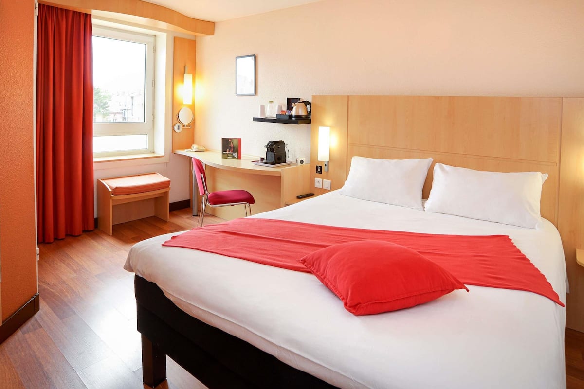 A hotel room with a bed, a desk, a chair, a window with curtains, a wall-mounted lamp, and a shelf with items.