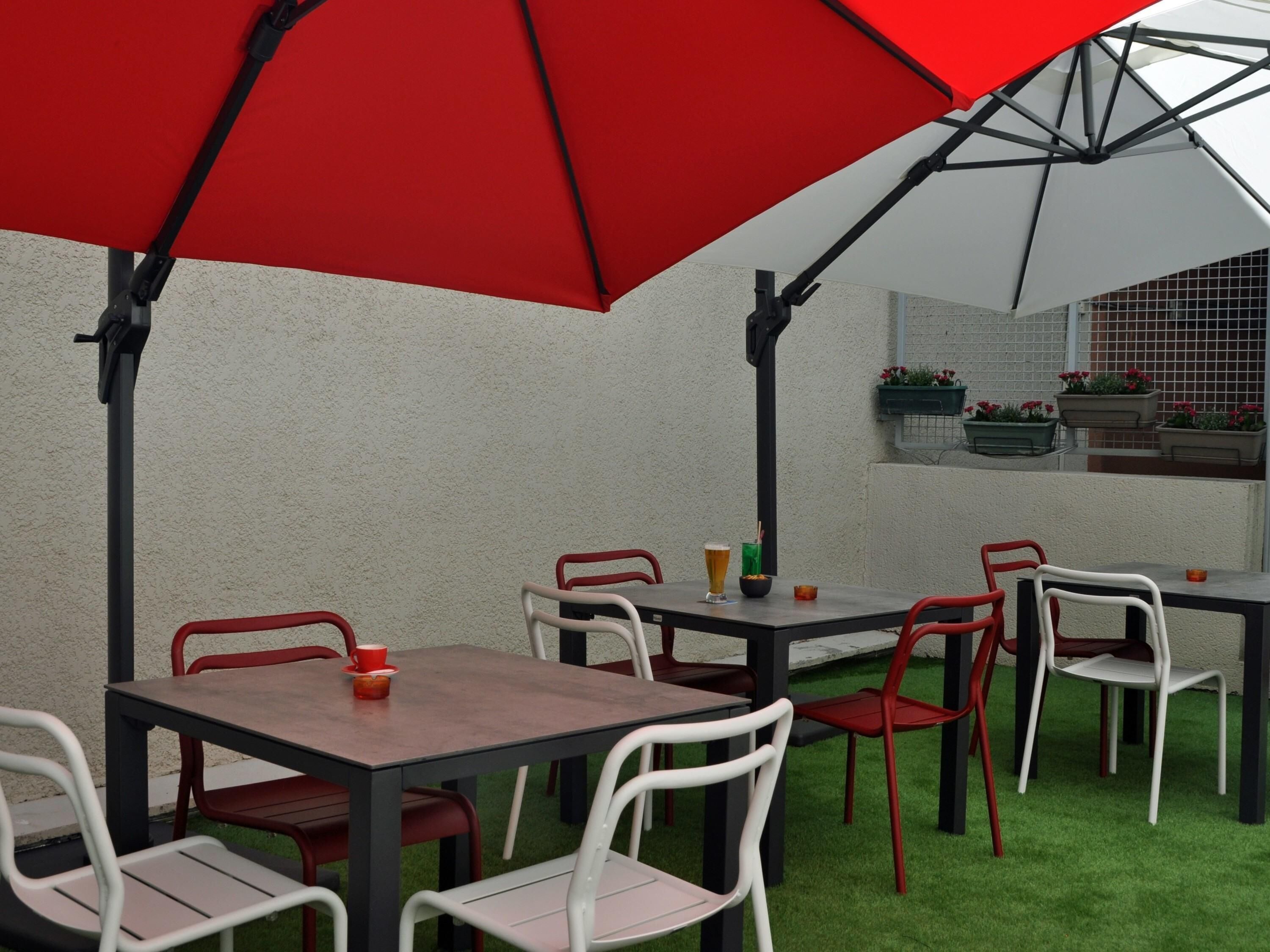 Un patio al aire libre con mesas y sillas, un paraguas rojo y vistas a un edificio con una ventana y jardineras.