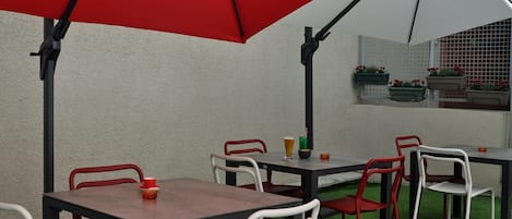 Un patio al aire libre con mesas y sillas, un paraguas rojo y vistas a un edificio con una ventana y jardineras.
