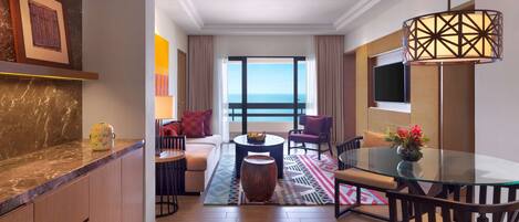 A modern hotel room with a marble countertop, a sofa with red cushions, a glass dining table, and a view of the ocean.
