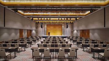 A conference room with rows of tables and chairs, a stage with a podium, and a large screen.