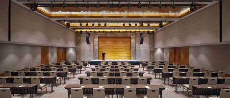 Une salle de conférence avec des rangées de tables et de chaises, une scène avec un pupitre et un grand écran.