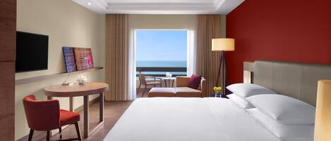 A hotel room with a large bed, a desk with a chair, a TV, and a view of the ocean.