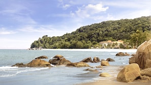 Una playa con rocas grandes, agua azul cristalina y una colina con árboles y edificios al fondo.