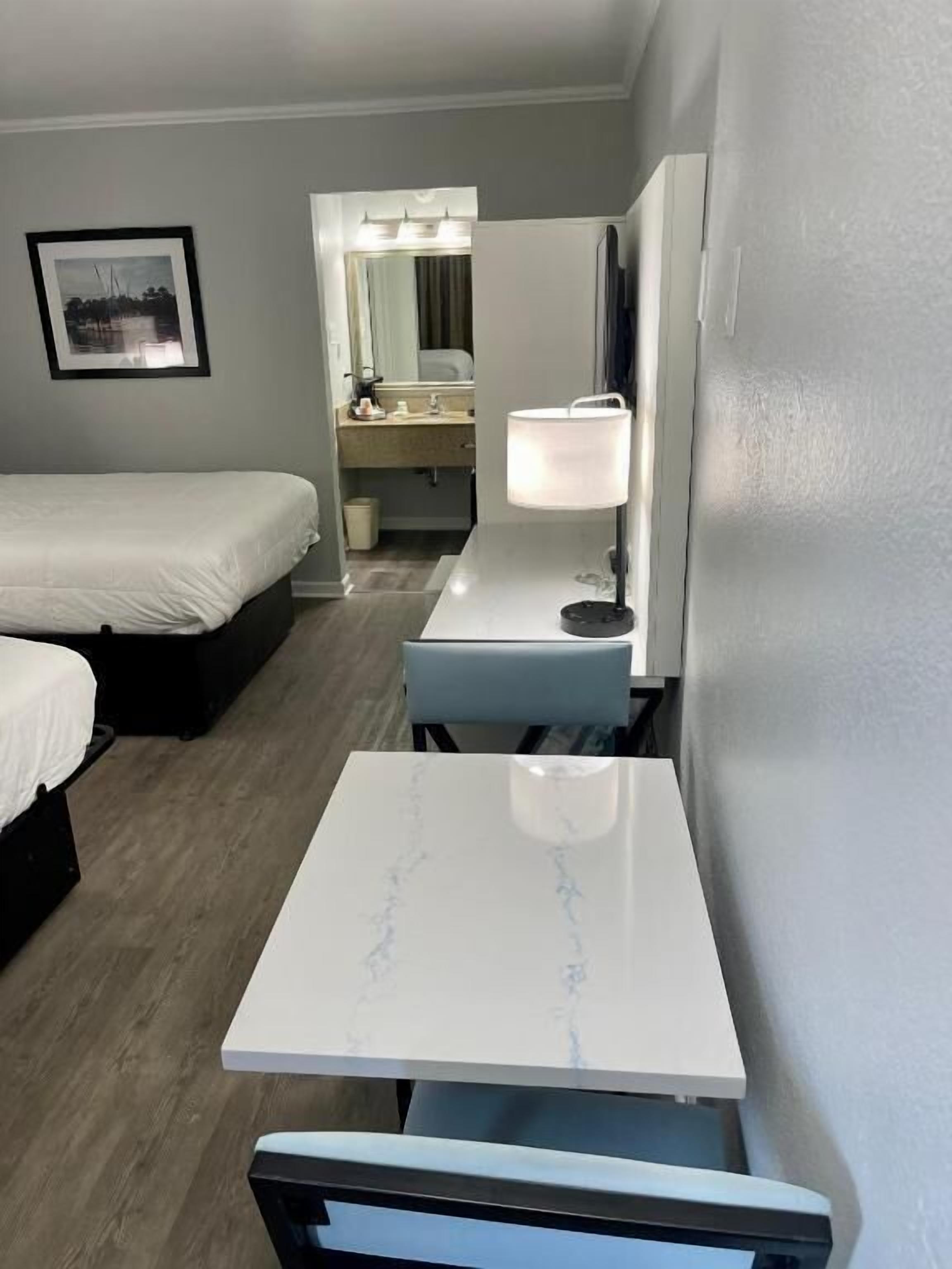 A hotel room with a bed, a desk with a lamp, a chair, a small table, and a bathroom visible in the background.