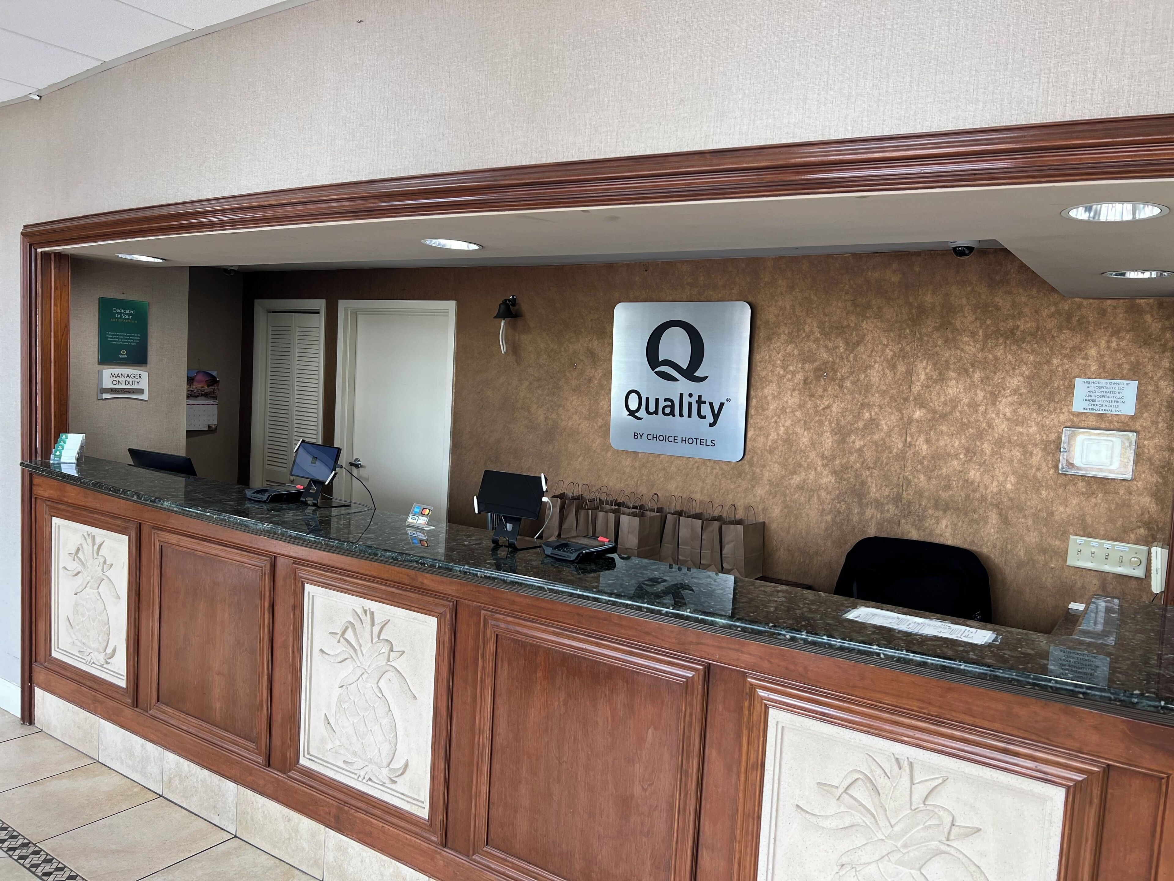 A hotel reception desk with a 'Quality by Choice Hotels' sign, a 'Manager on Duty' sign, and a security camera.