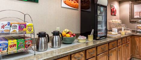 A hotel kitchen with cereal boxes, a fruit bowl, a coffee maker, and a refrigerator.