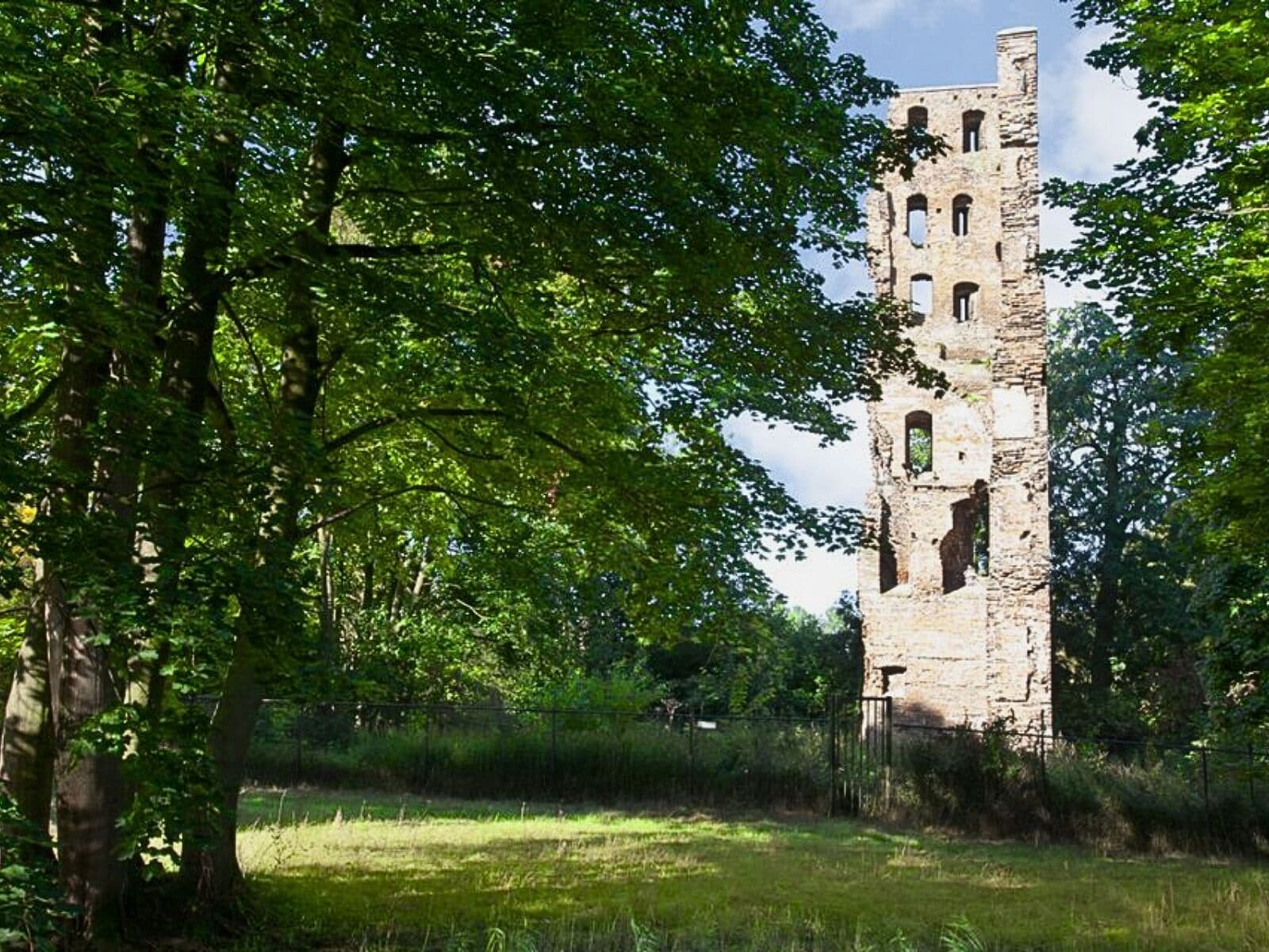 A historic stone tower with multiple arched windows, surrounded by lush green trees and grass.