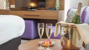 A hotel room with a bed, a TV, a minibar, a table with two glasses of champagne, a bottle of champagne, and a vase with flowers. - A-Hotel Oosterhout (Oosterhout)