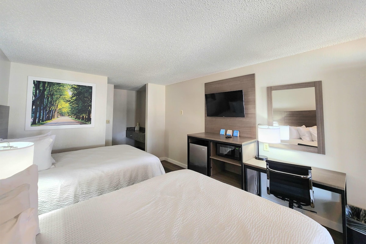 A hotel room with two beds, a desk with a computer, a TV, and a large window with a view of trees.