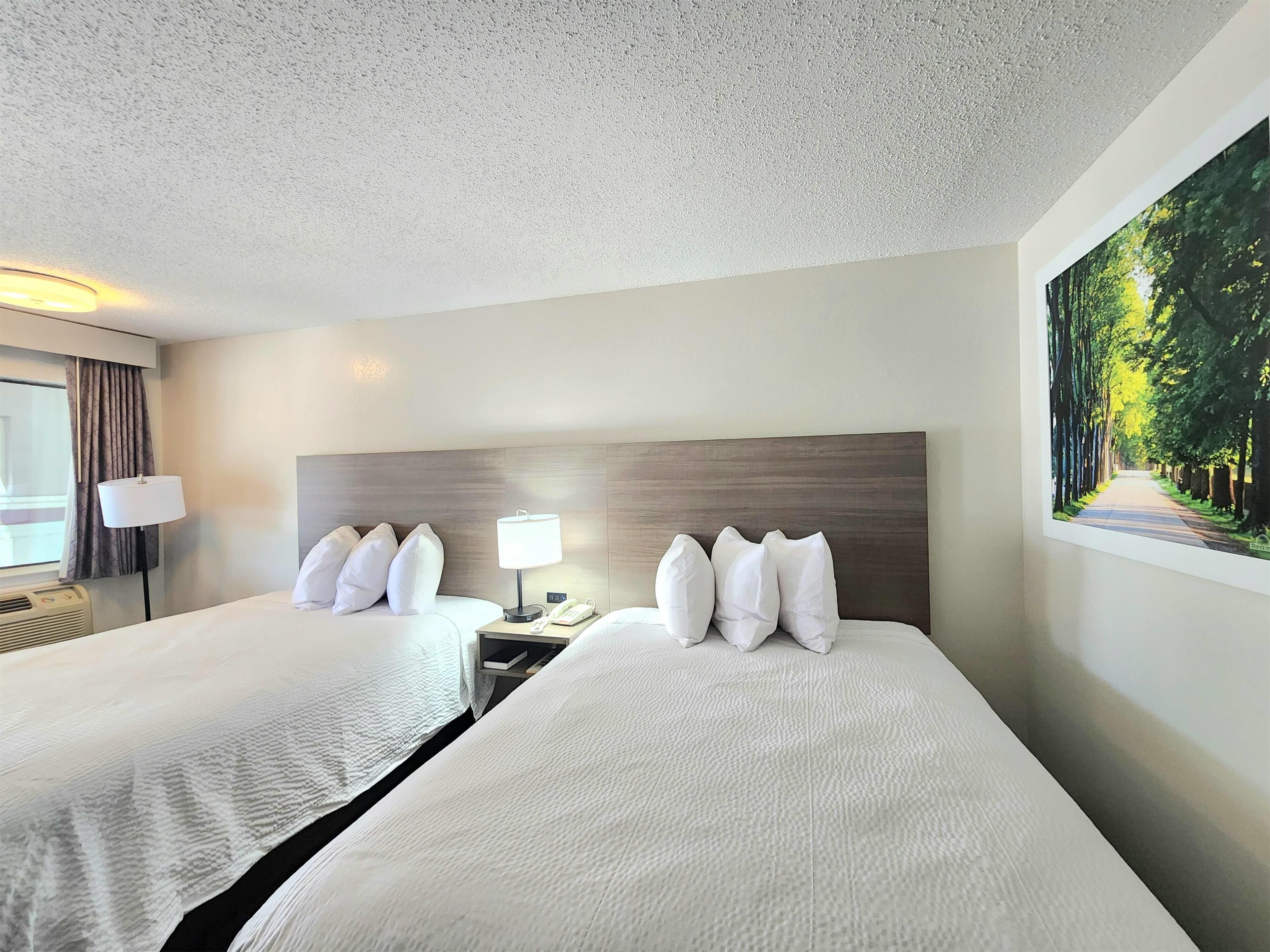 A hotel room with two beds, a nightstand with a lamp, a painting of a forest, and a window with curtains.