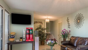 A living room with a brown leather couch, a flat-screen TV mounted on the wall, a coffee station with Folgers coffee and creamers, a water dispenser, and a potted plant with purple flowers. - Days Inn by Wyndham San Marcos (San Marcos)
