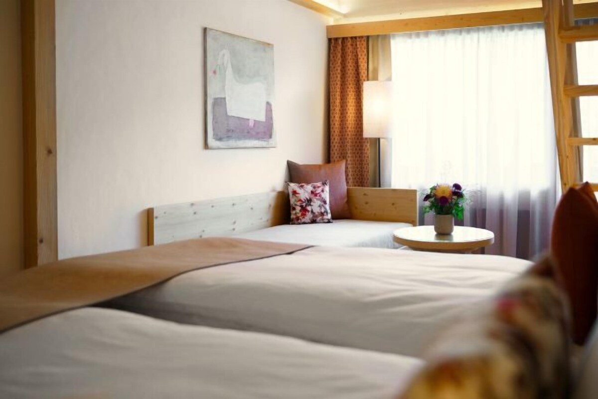 A hotel room with two beds, a small table with a vase of flowers, a painting on the wall, and a window with curtains.