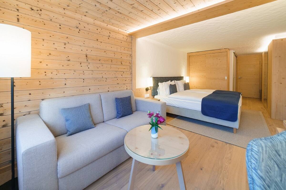 Moderni hotellihuone, jossa on puinen katto, sinisellä peitolla varustettu sänky, sinisillä koristetyynyillä varustettu sohva, pieni pyöreä pöytä, jossa on maljakko täynnä kukkia, ja yöpöytä, jossa on lamppu.