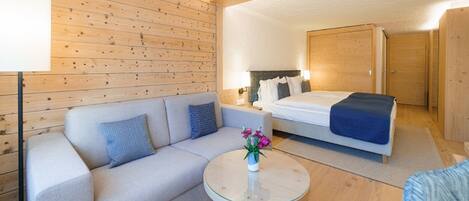 A modern hotel room with a wooden ceiling, a bed, a sofa, a small round table with a vase of flowers, and a nightstand with a lamp.