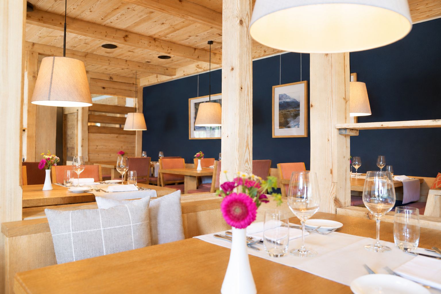 A restaurant interior with wooden tables and chairs, hanging pendant lights, and framed artwork on the walls.