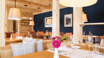 A restaurant interior with wooden tables and chairs, hanging pendant lights, and framed artwork on the walls.