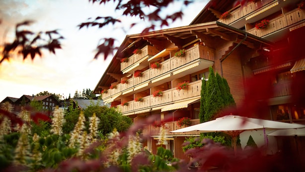 A multi-story wooden building with balconies adorned with red flowers, surrounded by lush greenery. - Gstaaderhof – Active & Relax Hotel  (Saanen)