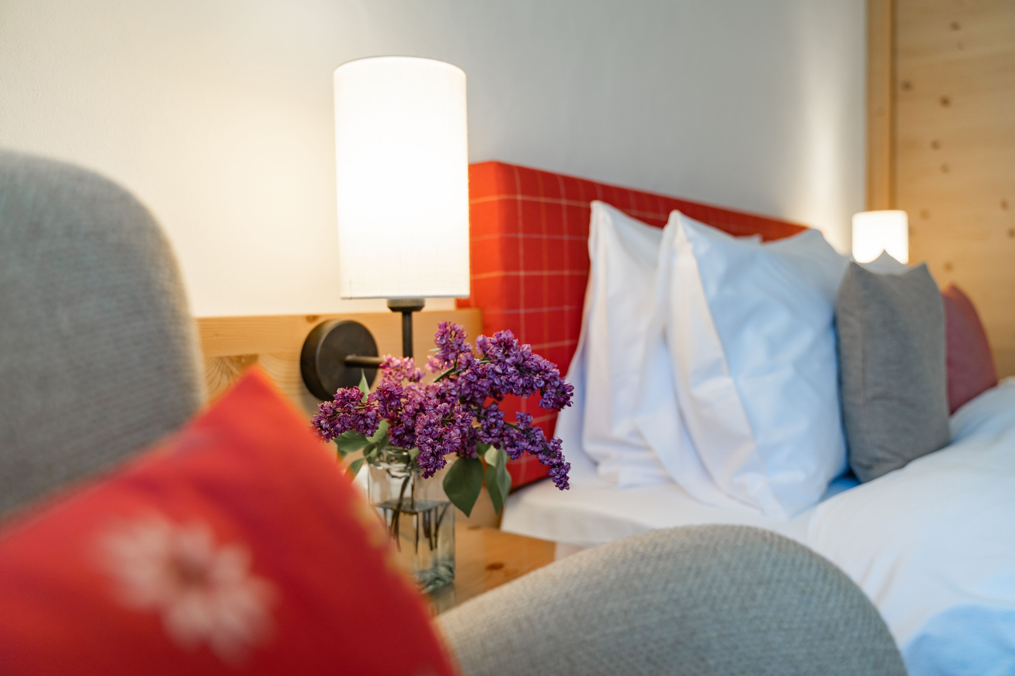 A hotel room with a bed, pillows, a bedside table with a lamp, and a vase with flowers.