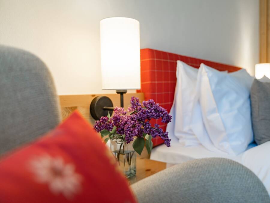A hotel room with a bed, pillows, a bedside table with a lamp, and a vase with flowers.