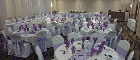 A banquet hall with round tables, each set with white tablecloths and purple sashes, and chairs with white covers and purple sashes.