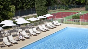 A swimming pool with sun loungers and umbrellas, surrounded by a tennis court and greenery. - Novotel Avignon Nord (Sorgues)