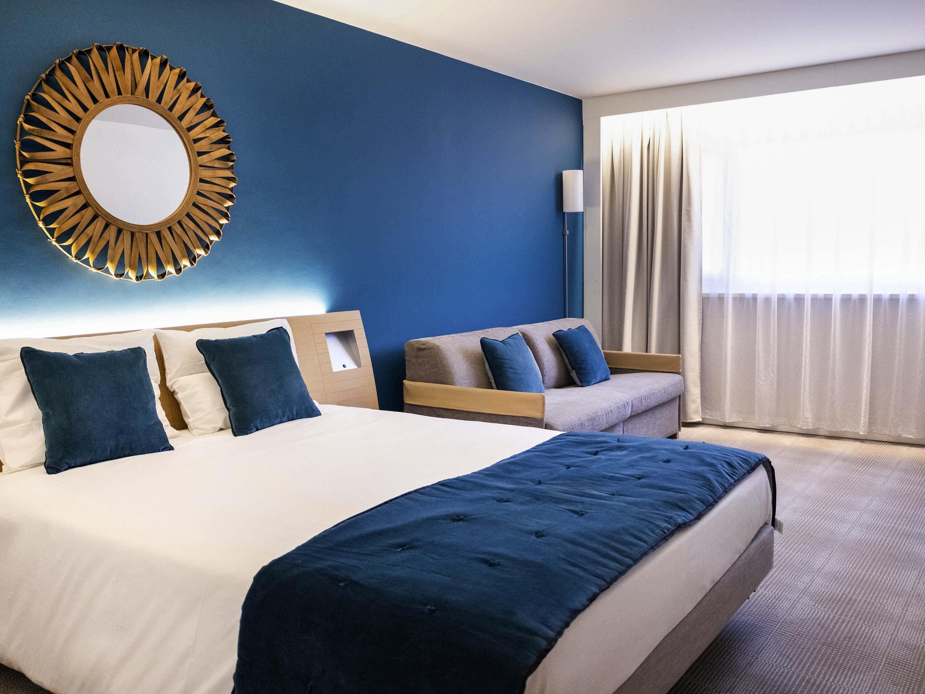 Et hotelværelse med en stor seng, en sofa, et spejl på væggen og et vindue med gardiner.