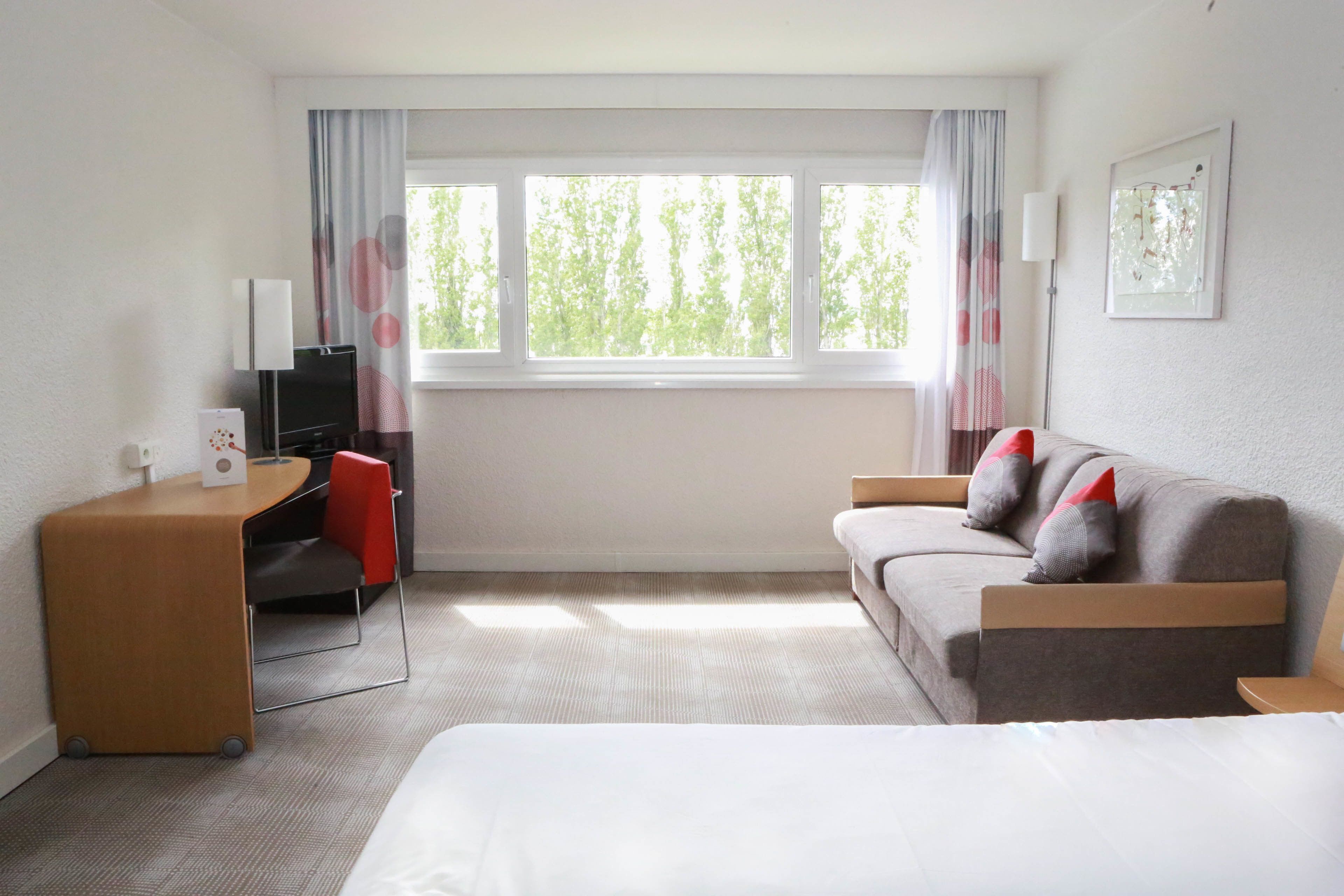 Habitació d'hotel amb llit, escriptori i cadira, sofà amb coixins, televisió i finestra amb cortines.