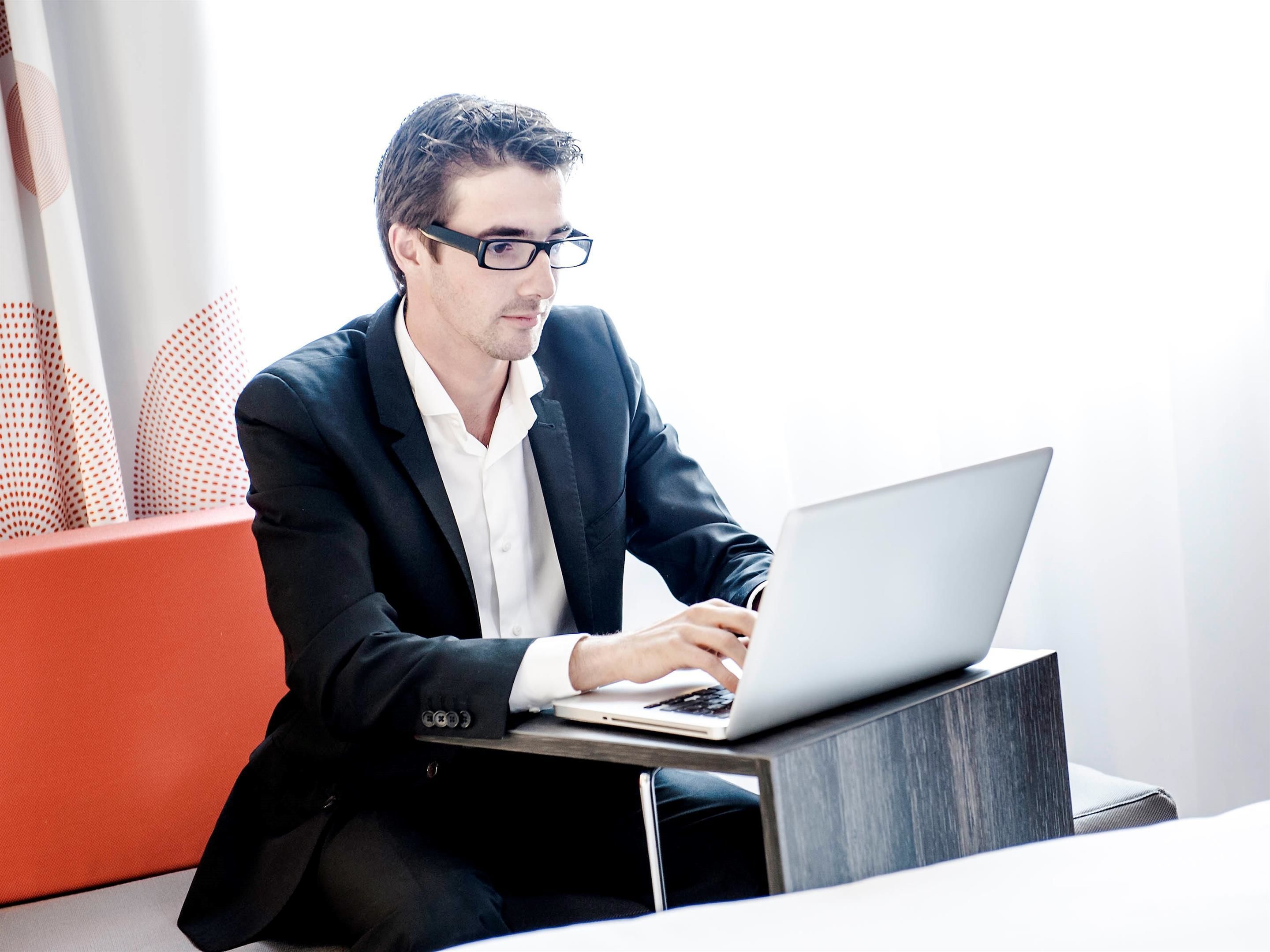 Un hombre con traje negro usando una computadora portátil sobre un escritorio moderno y minimalista en una habitación luminosa con una pared de acento roja.