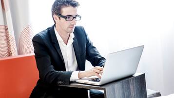 Un hombre con traje negro usando una computadora portátil sobre un escritorio moderno y minimalista en una habitación luminosa con una pared de acento roja.