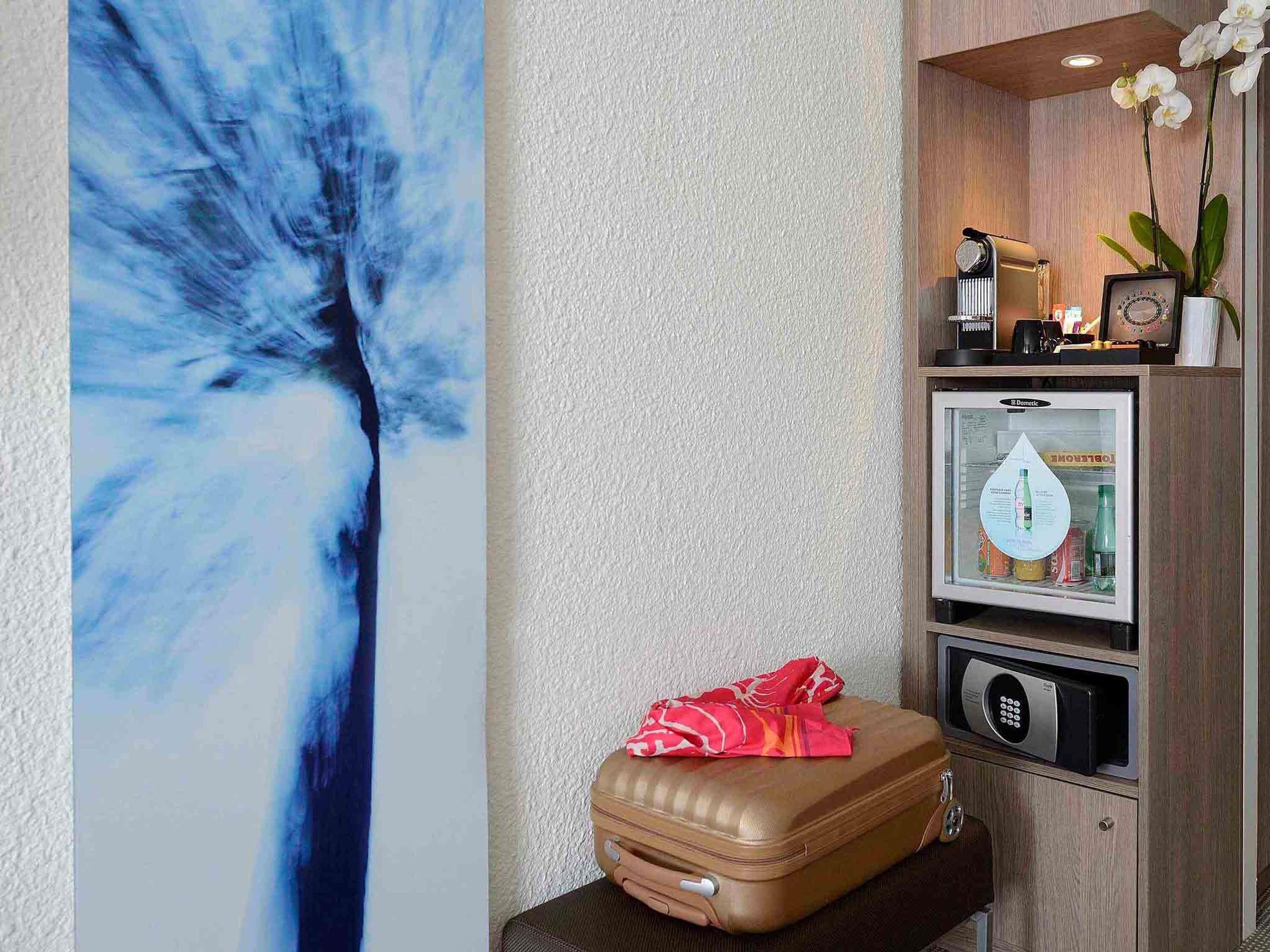 Una habitación con un cuadro abstracto grande, un minibar con refrigerador, una máquina de café, un reloj y una maleta sobre una mesa.