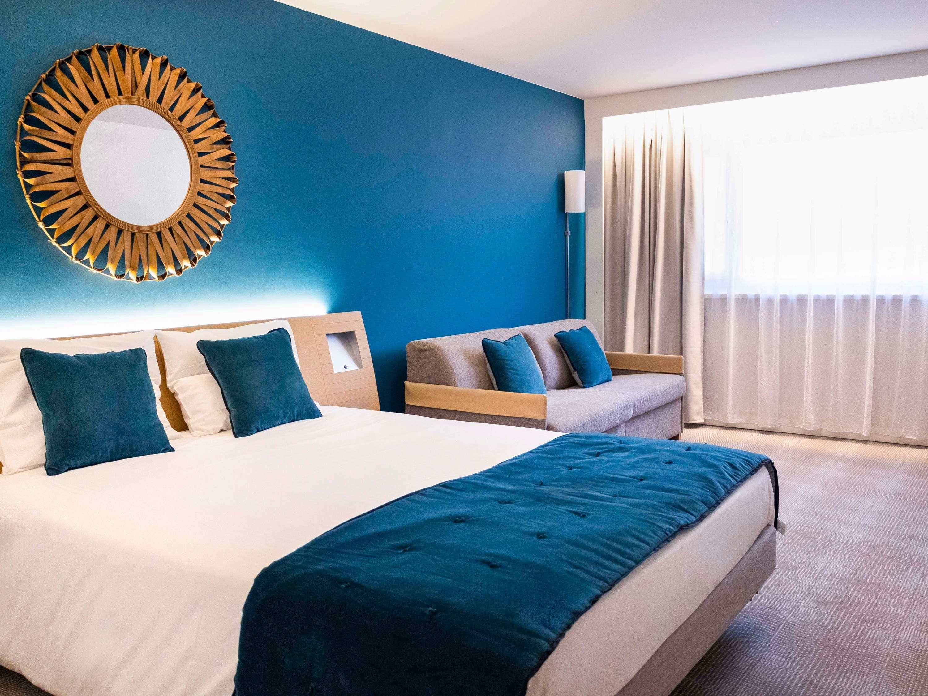 Una habitación de hotel con una cama grande, un sofá, un espejo montado en la pared y una ventana con cortinas.