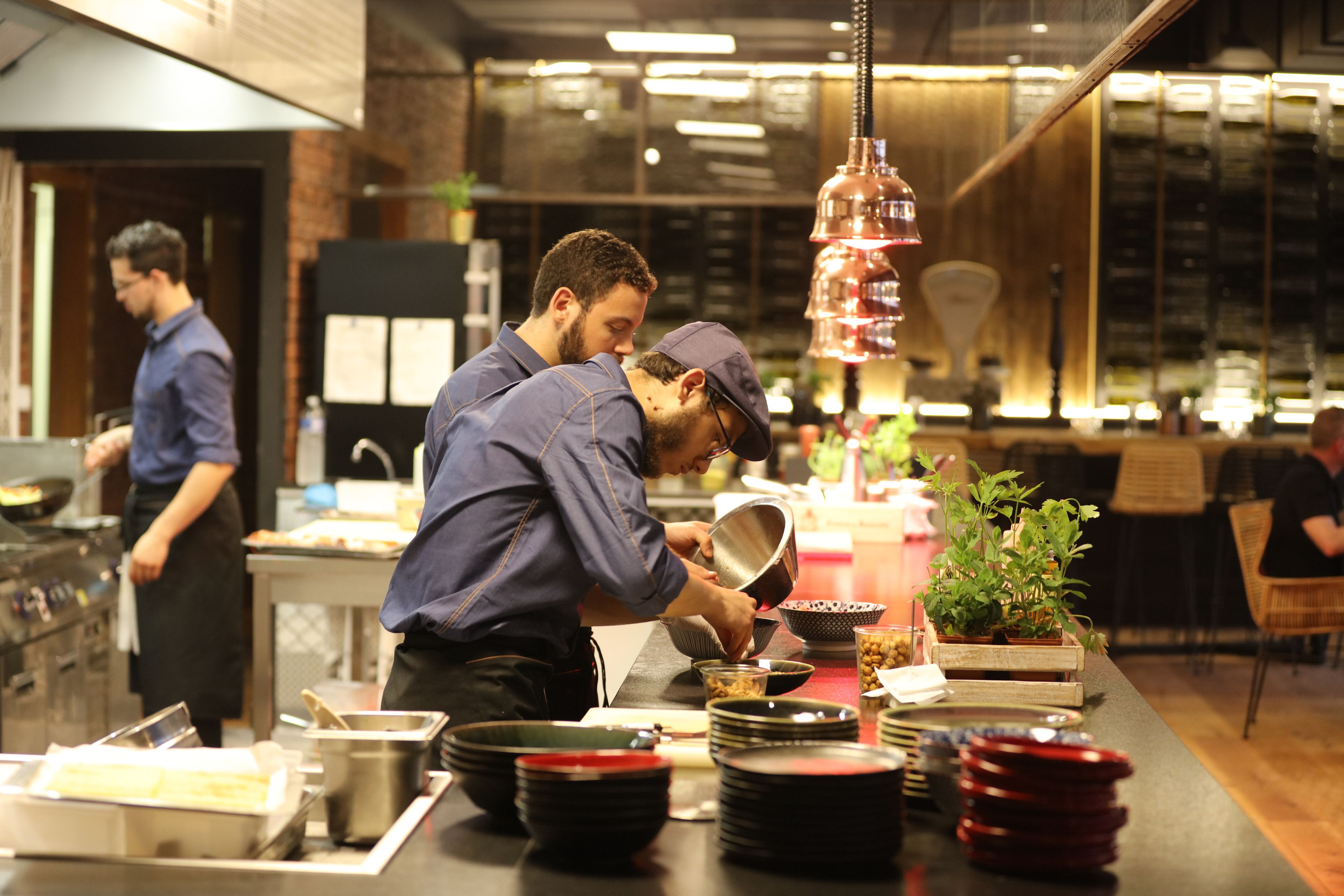 Una cocina de restaurante con cocineros preparando alimentos, una barra con platos apilados y una lámpara colgante.