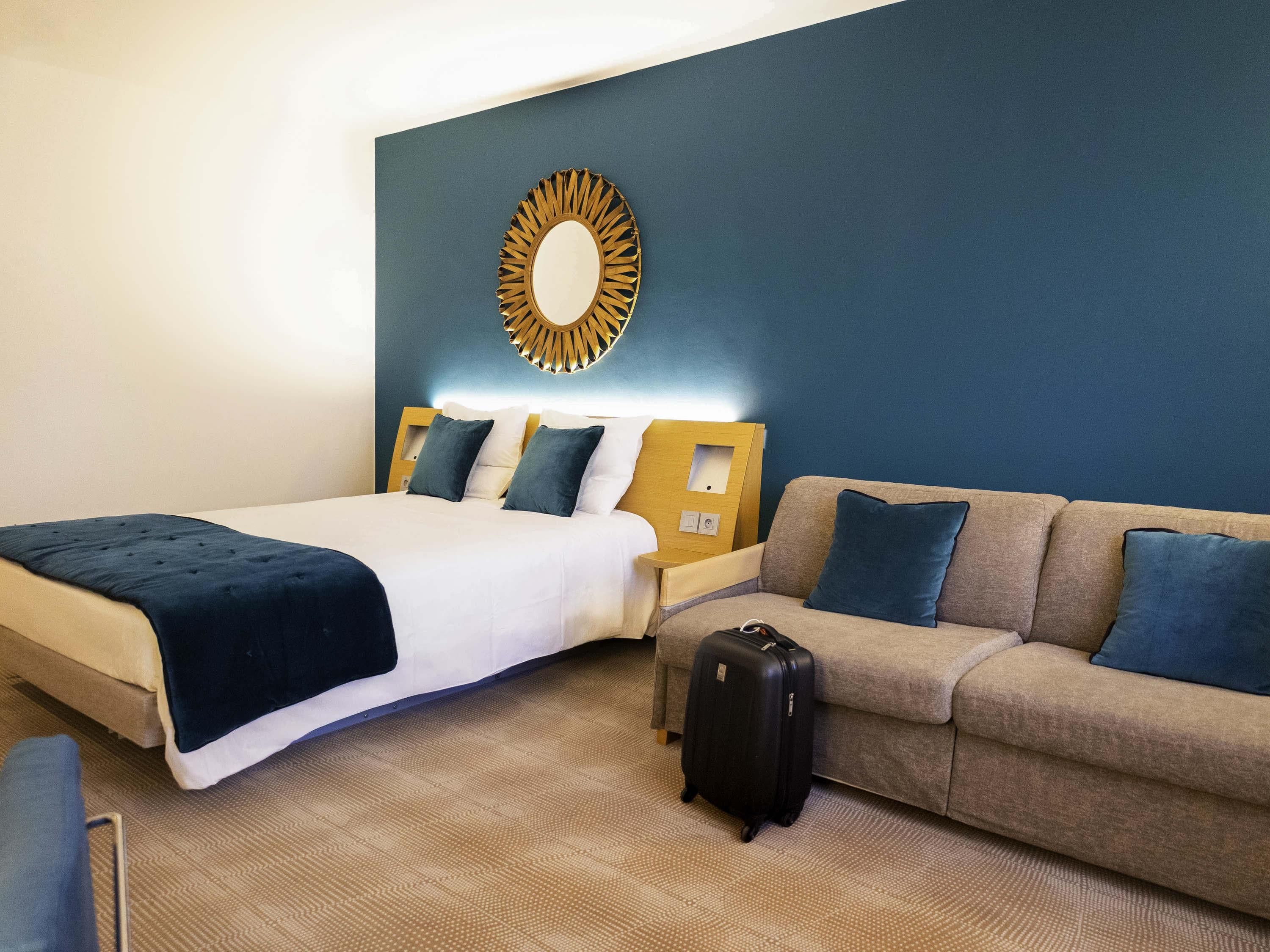 Una habitación de hotel con una cama, un sofá, una maleta y un espejo fijado a la pared.