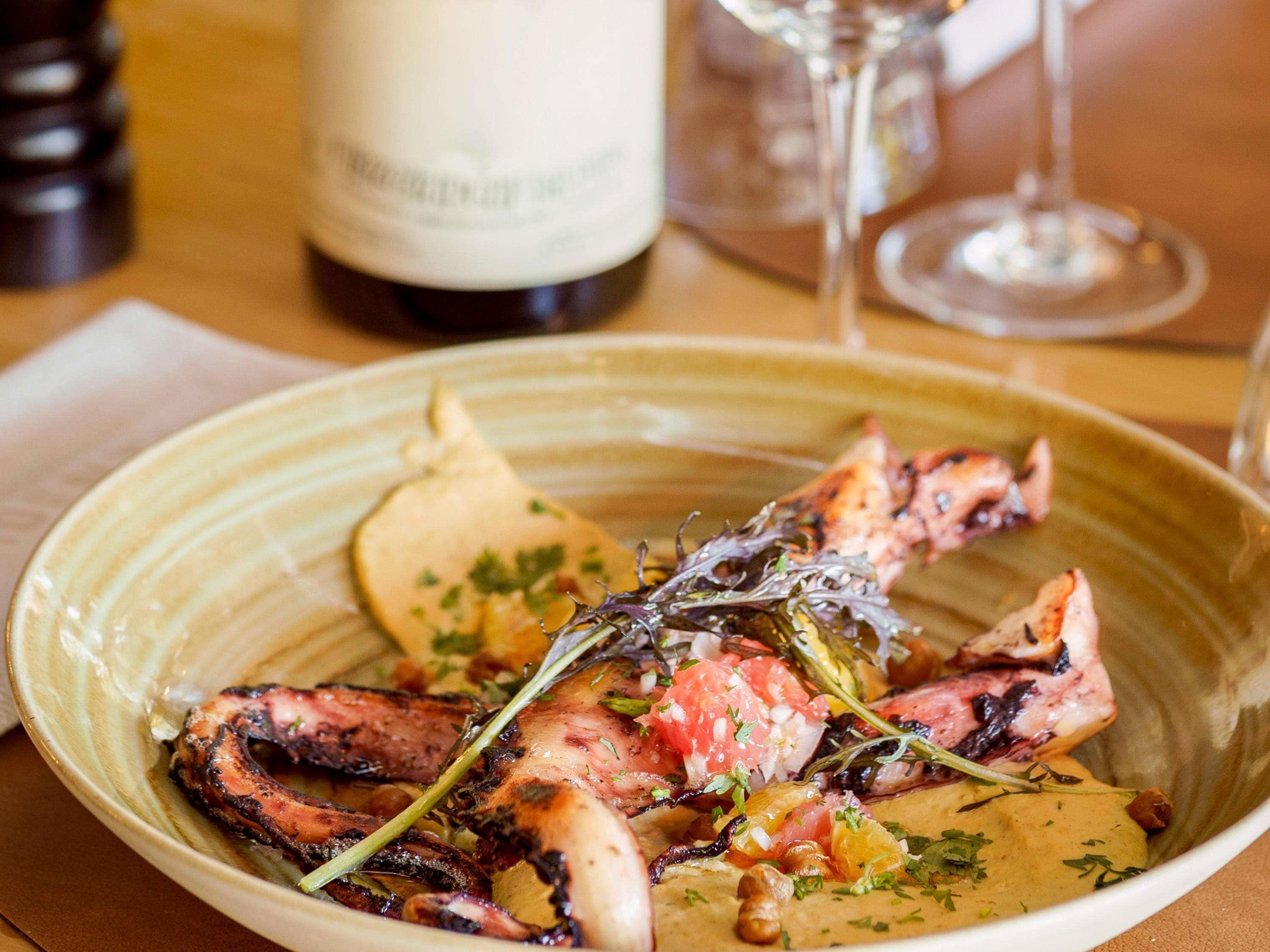 A plate of grilled octopus with vegetables and herbs, accompanied by a bottle of wine and a glass of wine on a table.