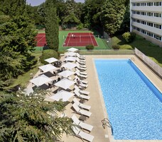 Piscina com espreguiçadeiras e guarda-sóis, quadra de tênis e um edifício ao fundo.
