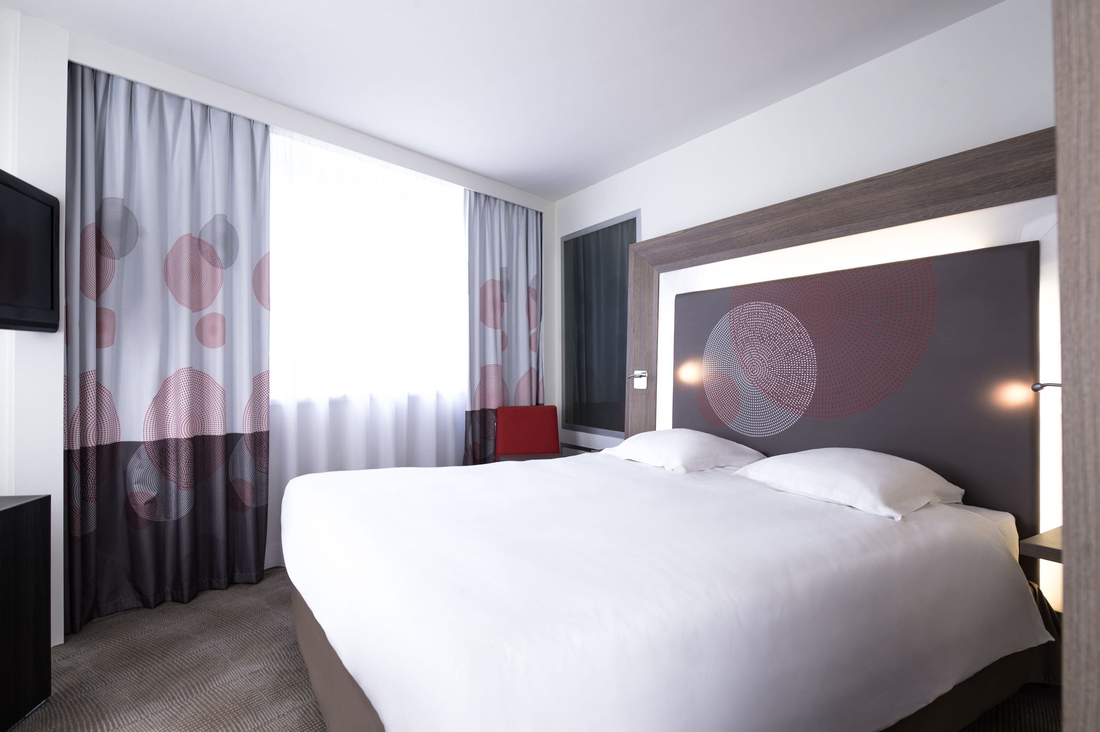 A modern hotel room with a large bed, a red chair, and a wall-mounted TV.